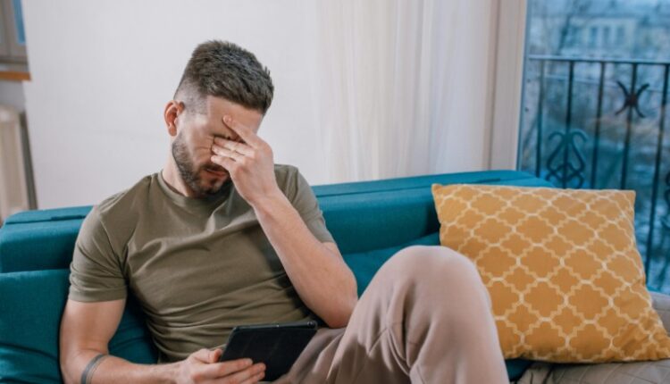 A man sits on a sofa holding a tablet, visibly stressed and covering his face with one hand., tags: online-casinos - pexel